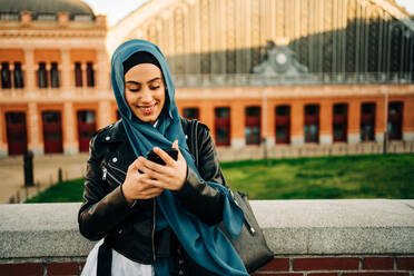 Muslimische Frau mit traditionellem Kopftuch steht auf einer Straße in der Stadt und surft auf ihrem Handy - ADSF26136