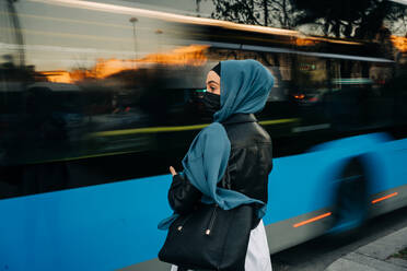 Rückenansicht einer Muslima im Hijab, die in der Nähe einer Straße mit regem Verkehr in der Hauptverkehrszeit steht - ADSF26141