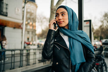 Charmante muslimische Frau mit Kopftuch steht auf der Straße und telefoniert, während sie wegschaut - ADSF26143