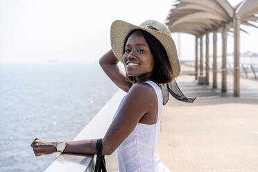 Seitenansicht afroamerikanische Frau in stilvollem weißem Kleid steht auf Stadtpromenade und genießt Meerblick auf sonniges Wetter - ADSF26145