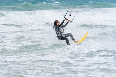 Aktive Sportlerin auf einem Kiteboard, die die Steuerstange hält, während sie Kitesurfen übt und auf den schaumigen Ozean blickt - ADSF26150