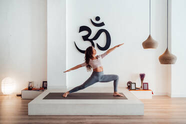Anmutige junge Frau steht in Virabhadrasana und übt Yoga auf der Matte im Studio - ADSF26159