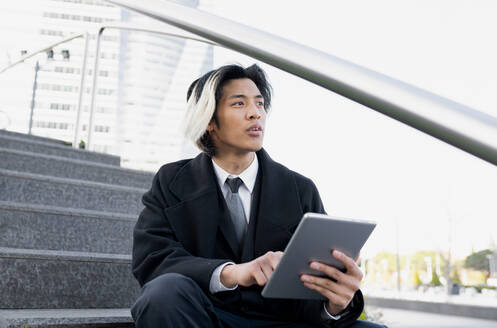 Junge, gut gekleidete männliche Führungskraft, die im Internet auf einem Tablet surft, während sie auf einer Treppe in der Stadt bei Tageslicht sitzt und wegschaut - ADSF26162