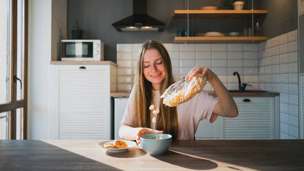 Fröhliche Frau schüttet knuspriges Müsli in eine Schüssel am Tisch mit leckeren Haferkeksen mit Schokoladenstückchen zum Frühstück zu Hause - ADSF26206