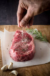 Crop anonymous male cook seasoning uncooked meat piece with salt and ground black pepper between garlic cloves and fresh rosemary sprig - ADSF26218