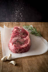 Raw beef piece with ground black pepper under salt in air between fresh garlic cloves and rosemary sprig - ADSF26221