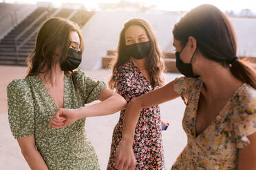 Content best female friends in ornamental dresses and cloth face masks touching elbows while looking at each other in town during coronavirus pandemic - ADSF26228