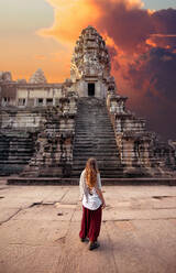 Back view of unrecognizable female tourist admiring old masonry worship exterior with staircase under cloudy sky at sundown in Cambodia - ADSF26231