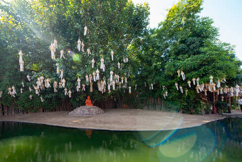 Buddha-Skulptur auf Stein unter Laternen auf Bäumen gegen orientalische Konstruktion reflektierend in reinem Teich in Thailand - ADSF26233