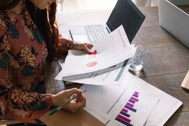 Crop fokussiert weiblich in stilvollem Kleid Büroangestellte Analyse von Diagrammen beim Sitzen am Schreibtisch mit Laptop und Papiere - ADSF26263