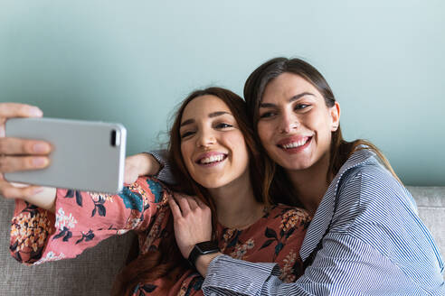 Junge glückliche weibliche Freunde in lässigen Outfits, die sich umarmen und ein Selfie mit dem Handy im hellen Wohnzimmer machen - ADSF26274