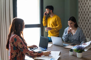 Group of young coworkers in casual clothes gathering around desk documents while working on netbooks and talking on smartphone - ADSF26276