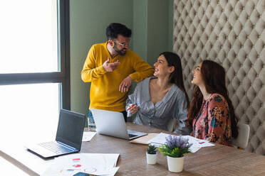 Eine Gruppe fröhlich lachender Mitarbeiter in legerer Kleidung versammelt sich am Schreibtisch mit Laptops und bespricht geschäftliche Details, während sie in einem hellen, modernen Büro zusammenarbeiten - ADSF26278