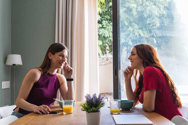 Fröhliche junge Freundinnen in legerer Kleidung, die zusammen lecker zu Mittag essen und ihr Smartphone benutzen, während sie am Esstisch in einer modernen Wohnung sitzen und sich unterhalten - ADSF26281
