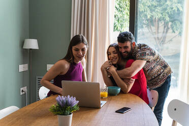 Gruppe junger, fröhlicher Freunde in Freizeitkleidung, die sich umarmen und am Esstisch versammeln, während sie auf einem modernen Netbook surfen und lächeln - ADSF26291
