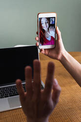 Crop-Frau winkt mit der Hand und grüßt ihre Freundin, während sie einen Videoanruf über ein Mobiltelefon führt und mit einem Laptop am Schreibtisch sitzt - ADSF26292