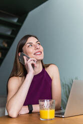Junge zufriedene Frau in Freizeitkleidung mit einem Mobiltelefon und am Tisch sitzend mit einem modernen Netbook in einem hellen Wohnzimmer - ADSF26295