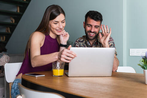 Zufriedenes junges Paar in legerer Kleidung, das gemeinsam auf einem modernen Netbook surft, während es am Tisch mit einem Glas Orangensaft im Wohnzimmer sitzt - ADSF26297