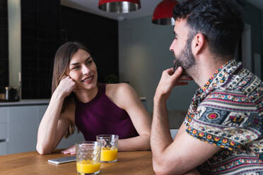 Positives junges Paar in Freizeitkleidung, das sich unterhält und frischen Orangensaft genießt, während es am Esstisch in der Küche sitzt - ADSF26298