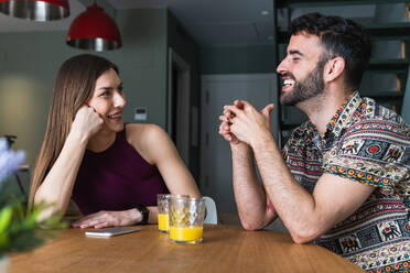 Positives junges Paar in Freizeitkleidung, das sich unterhält und frischen Orangensaft genießt, während es am Esstisch in der Küche sitzt - ADSF26299