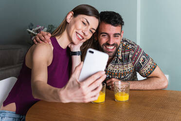 Fröhliches junges Paar in Freizeitkleidung, das sich umarmt und ein Selfie mit dem Handy macht, während es am Tisch mit Gläsern mit frischem Orangensaft sitzt - ADSF26300