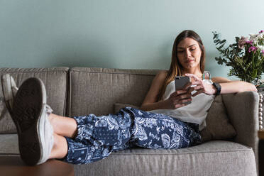 Adult female text messaging on cellphone while sitting with crossed legs on couch in living room - ADSF26301