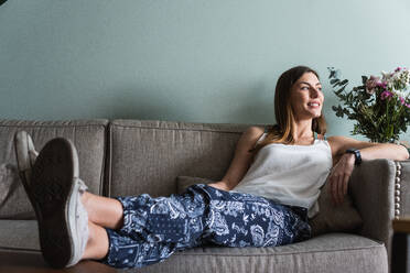 Kontemplative Frau in Hose mit Ornament sitzt mit gekreuzten Beinen auf der Couch und schaut nach Hause - ADSF26302