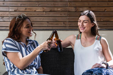 Lächelnde beste Freundinnen, die auf Flaschen mit alkoholischen Getränken anstoßen, während sie sich gegenseitig auf der Couch im Sonnenlicht anschauen - ADSF26305