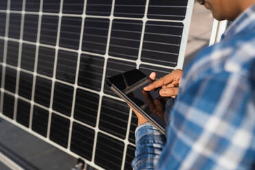 Abgeschnittener, nicht erkennbarer männlicher Techniker in kariertem Hemd, der in der Nähe eines Photovoltaik-Paneels in einem modernen Solarkraftwerk steht und ein Tablet betrachtet - ADSF26329