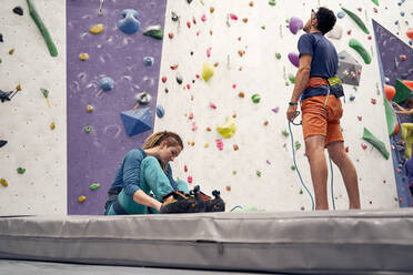 Ein männlicher Ausbilder mit Klettergurt und eine Frau, die auf dem Boden sitzt und sich auf das Klettern im Boulder-Club vorbereitet, von unten - ADSF26331