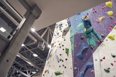 Von unten von einer starken Frau in einem Sicherheitsgurt beim Klettern an der Wand während des Trainings in einer Boulderhalle - ADSF26333