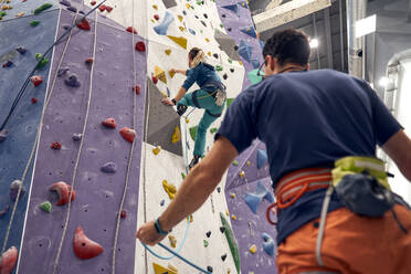 Von unten: mutige Sportlerin beim Klettern an der künstlichen Wand im Bouldering Center unter Aufsicht eines professionellen Trainers - ADSF26336