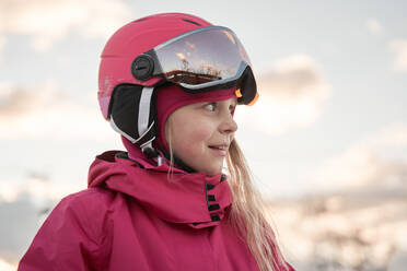 Fröhliches, liebenswertes Mädchen in rosa, warmer Sportkleidung und Helm, das auf einem weitläufigen, verschneiten Gelände steht und wegschaut - ADSF26348