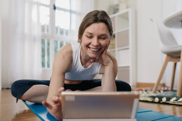 Delighted slim female sitting on mat and browsing tablet while choosing online lesson for practicing yoga - ADSF26357