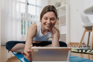 Zufriedene schlanke Frau, die auf einer Matte sitzt und auf einem Tablet surft, während sie eine Online-Lektion zum Üben von Yoga auswählt - ADSF26357