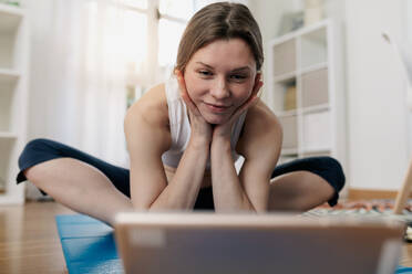 Delighted slim female sitting on mat and browsing tablet while choosing online lesson for practicing yoga - ADSF26358