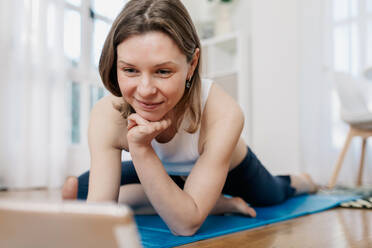 Delighted slim female lying on mat and browsing tablet while choosing online lesson for practicing yoga - ADSF26359