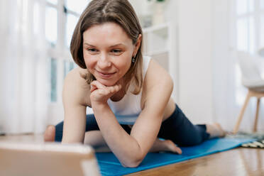 Zufriedene schlanke Frau, die auf einer Matte liegt und auf einem Tablet surft, während sie eine Online-Lektion zum Üben von Yoga auswählt - ADSF26359