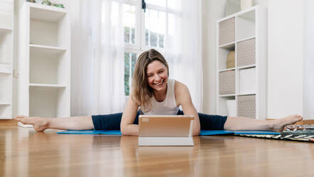 Lächelnde, flexible Frau, die Yoga in Upavista Konasana übt, während sie auf einer Matte sitzt und ein Online-Video-Tutorial auf einem Tablet zu Hause anschaut - ADSF26360