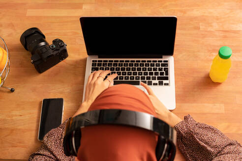 Von oben anonyme Frau mit Kopftuch, die an einem Schreibtisch sitzend ein modernes Netbook mit schwarzem Bildschirm benutzt - ADSF26380