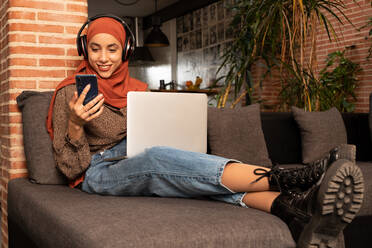 Voller Länge glücklich junge muslimische Frau in bescheidener Kleidung Hijab und drahtlose Kopfhörer sitzen auf gemütliche Couch mit Netbook auf dem Schoß und Surfen Smartphone - ADSF26382