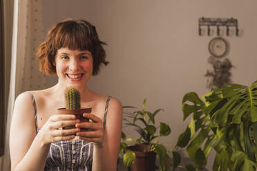 Happy woman with brown hair holding cactus plant at home - MGRF00335