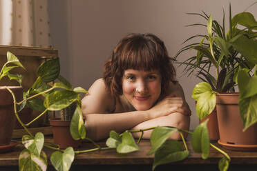Lächelnde junge Frau, die sich zu Hause an einen Tisch mit Pflanzen lehnt - MGRF00338