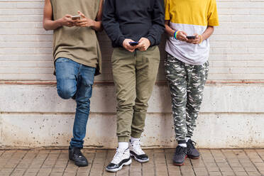 Crop diverse teens addicted to mobile phones browsing Internet while leaning on brick wall - ADSF26386