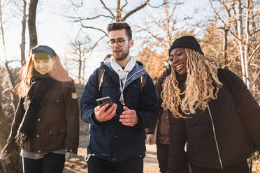 Multiethnische Freunde in Oberbekleidung stehen im Kreis und teilen sich ein Mobiltelefon im Wald - ADSF26392