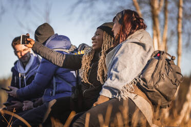 Glückliche junge gemischtrassige Freunde in warmer Kleidung sitzen auf einem Holzsteg mit Smartphones im See während einer Wanderung im Herbstwald an einem sonnigen Tag - ADSF26394