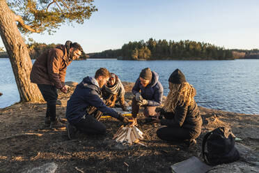 Full body of joyful young diverse travelling friends in warm clothes sitting on lake shore and lighting fire during camping in autumn forest on sunny day - ADSF26396