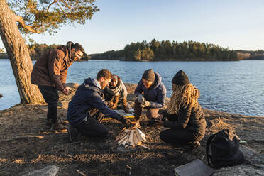 Ganzer Körper von fröhlichen jungen Reisenden in warmer Kleidung am Seeufer sitzend und Feuer anzündend beim Camping im Herbstwald an einem sonnigen Tag - ADSF26396
