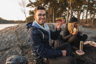 Fröhlicher junger männlicher Reisender in warmer Kleidung und mit Brille, der lächelnd ein leckeres Sandwich isst, während er mit einer afroamerikanischen Freundin in der Nähe eines Sees zeltet - ADSF26397