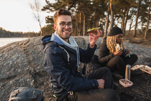 Cheerful young male traveler in warm clothes and eyeglasses smiling while eating tasty sandwich during camping with African American female friend near lake - ADSF26397